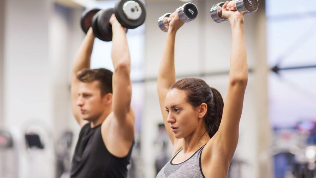 man and a woman perform standing dumbbell overhead presses near each other.