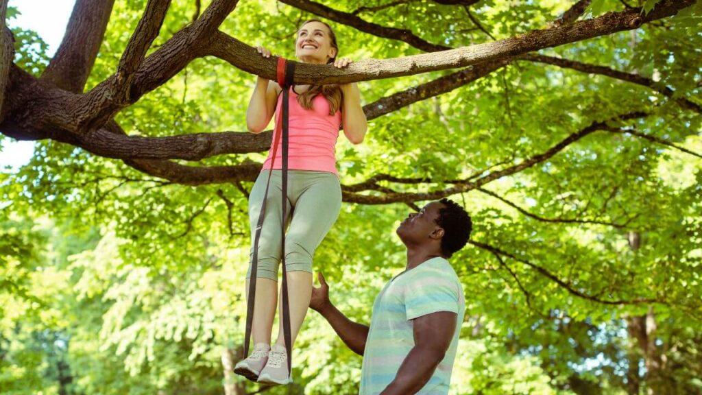 A man helps a woman perform assisted pull ups with resistance bands on the tree. This is the proper demonstration of how to use assistance bands for pull-ups.