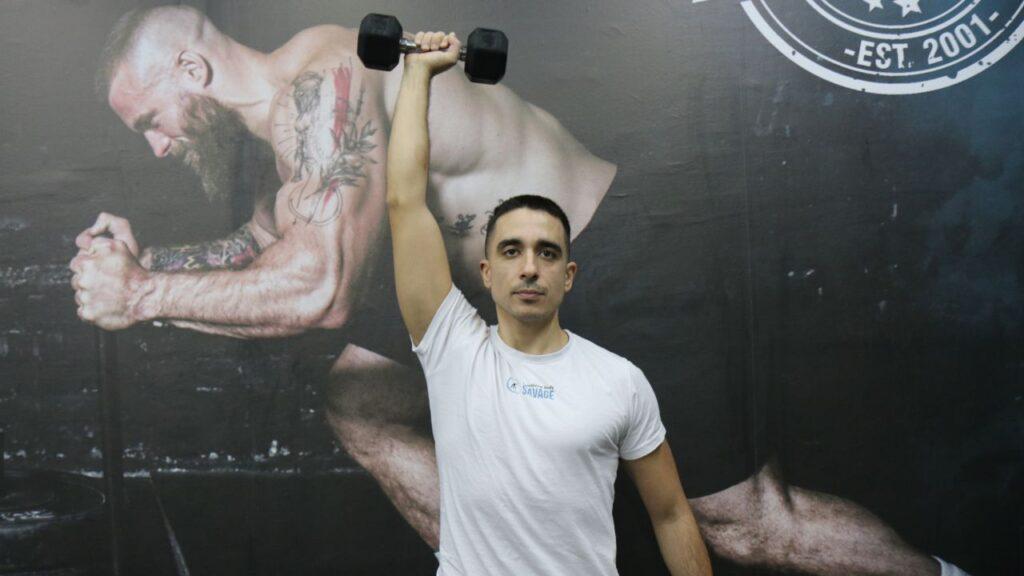 Radomir performs standing single-arm overhead dumbbell tricep extensions.
