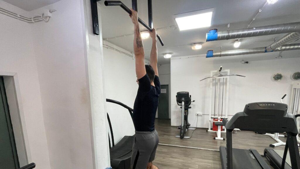 Vanja performs a dead hang exercise on the pull-up bar in the commercial gym setup.