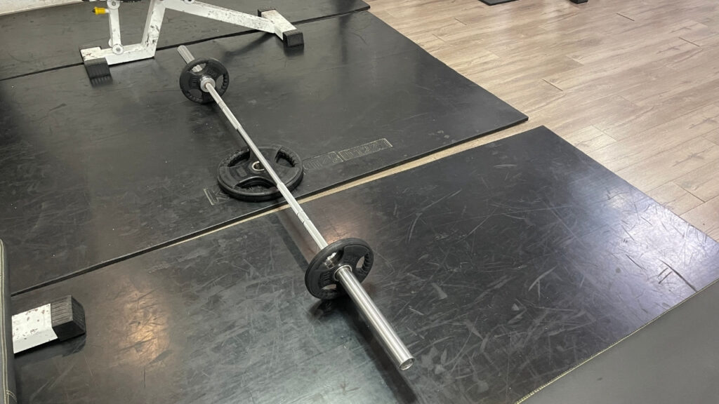 The image shows the first step of performing toes elevated RDLs. This step includes putting the barbell and a weight plate on the floor.