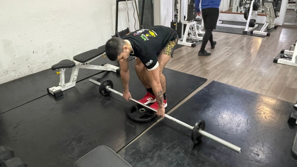 The image shows the second step of performing toes elevated RDLs. This step includes assuming the proper starting position in front of the barbell with your toes elevated on the weight plate.
